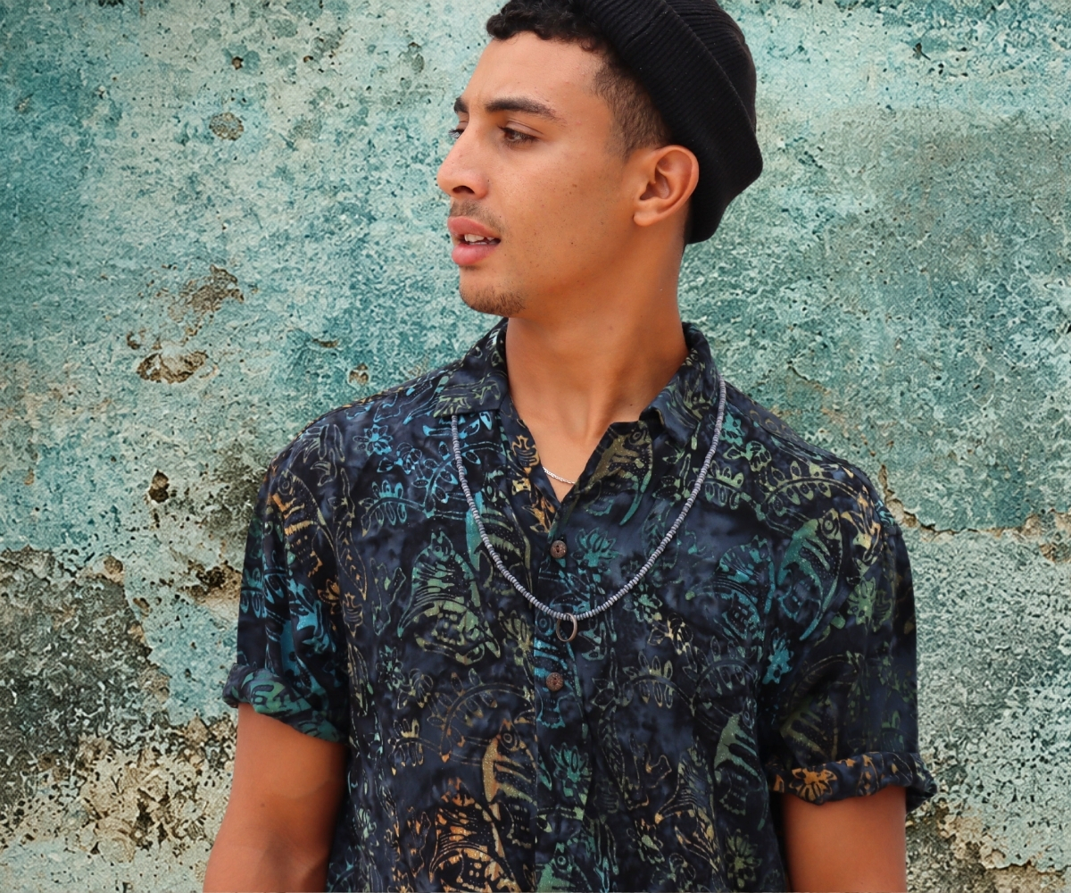 Man wearing a dark blue men’s short-sleeve shirt with batik pattern and black beanie against a textured wall