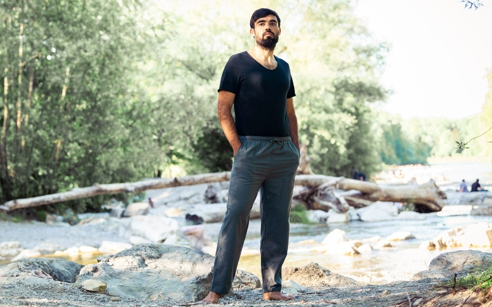 Mann trägt schwarzes Herren T-Shirt und graue Freizeithose, barfuß auf Steinen am Flussufer in natürlicher Outdoor-Umgebung