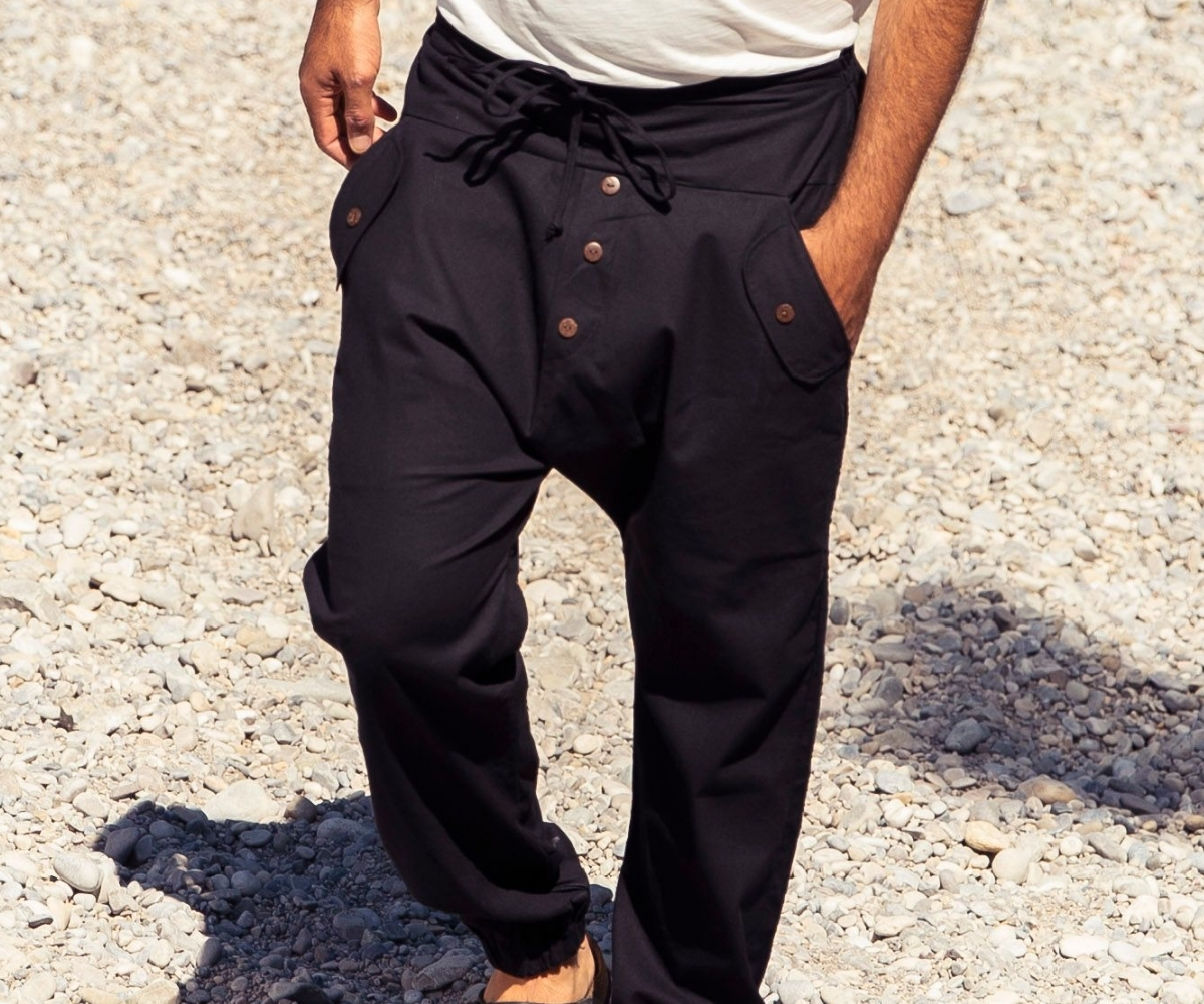 Man wearing black men's harem trousers with a low crotch, button placket and side pockets on stony ground