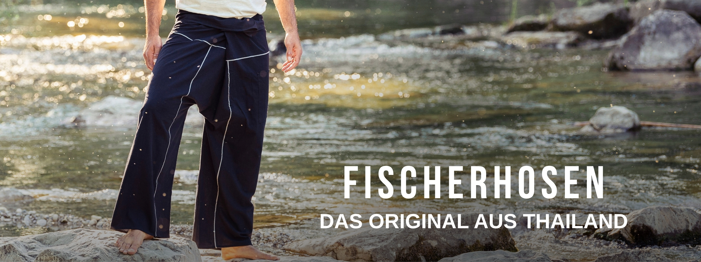 Model trägt eine dunkle Fischerhose aus Thailand und steht barfuß auf Felsen am Fluss