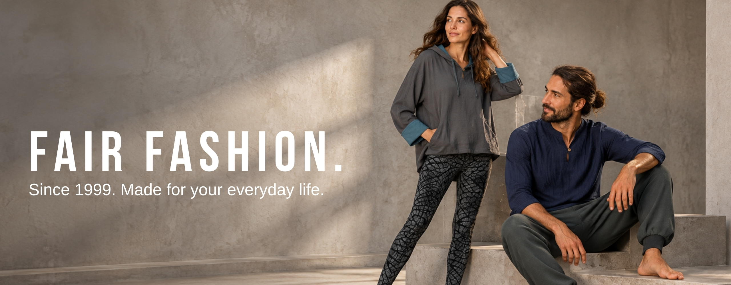 Woman and man in sustainable fair fashion loungewear against a concrete wall, wearing a hoodie, patterned leggings and a blue top