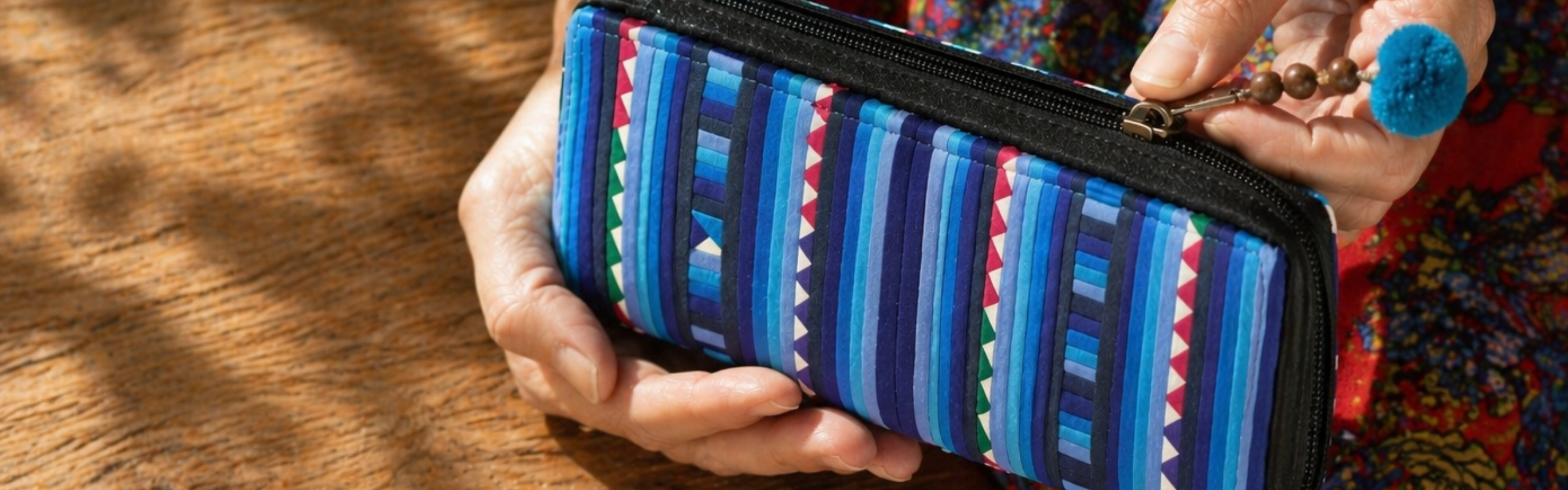Hands holding a colourful women's zip wallet with blue striped pattern and pom-pom charm